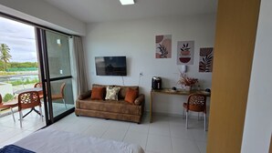 A modern hotel room with a glass sliding door leading to a balcony, a sofa with cushions, a small desk with a kettle, and wall art.