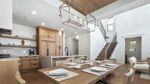 Una cocina amplia con armarios de madera, un comedor con una gran mesa de madera dispuesta para seis personas, y una escalera que lleva a un nivel superior.