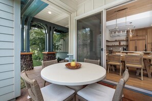 Une terrasse couverte avec une table ronde et des chaises en osier, un bac à fleurs en pierre et une vue sur une cuisine dotée de meubles en bois et d’un luminaire suspendu.