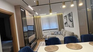 Uma sala de estar moderna com área de jantar, apresentando um conjunto de sofá, uma mesa de centro e uma janela grande com cortinas.