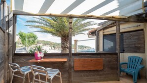 An outdoor patio with metal bar stools, a wooden table, a blue chair, and a view of a beach and palm tree. - Sable entre les Orteils - Studio vue mer (Banyuls-sur-Mer)
