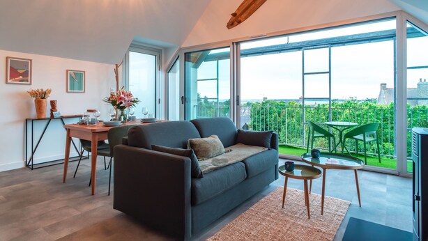 A modern living room with a sofa, dining table, and chairs, large windows with a view of greenery and buildings.