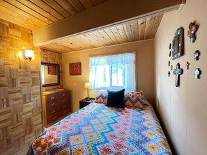Een slaapkamer met een houten plafond, een bed met een kleurrijke quilt, een dressoir, een nachtkastje met een lamp en decoraties aan de muur.