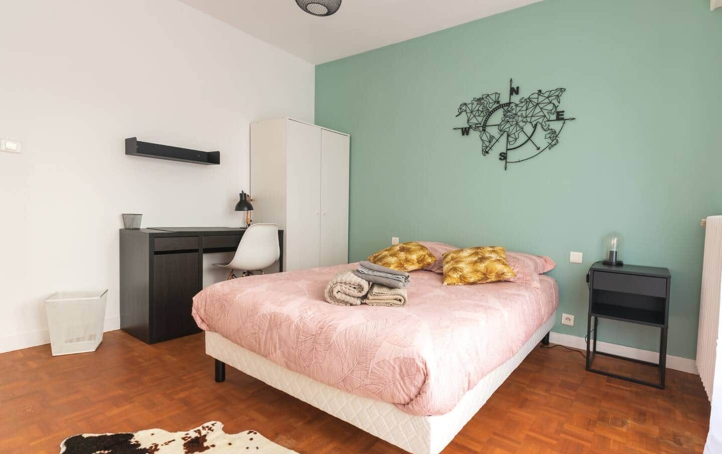 A bedroom with a bed, a desk, a chair, a wardrobe, a small table, and a wall art piece.