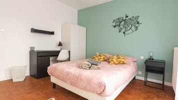 A bedroom with a bed, a desk, a chair, a wardrobe, a small table, and a wall art piece.