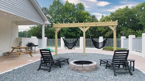 En udendørs terrasse med et træbeklædt pergola, to sorte hængekøjer, et bålsted og en grill.
