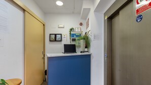 Una reception con un bancone blu, una porta chiusa, una pianta, certificati incorniciati e un orologio appeso al muro.