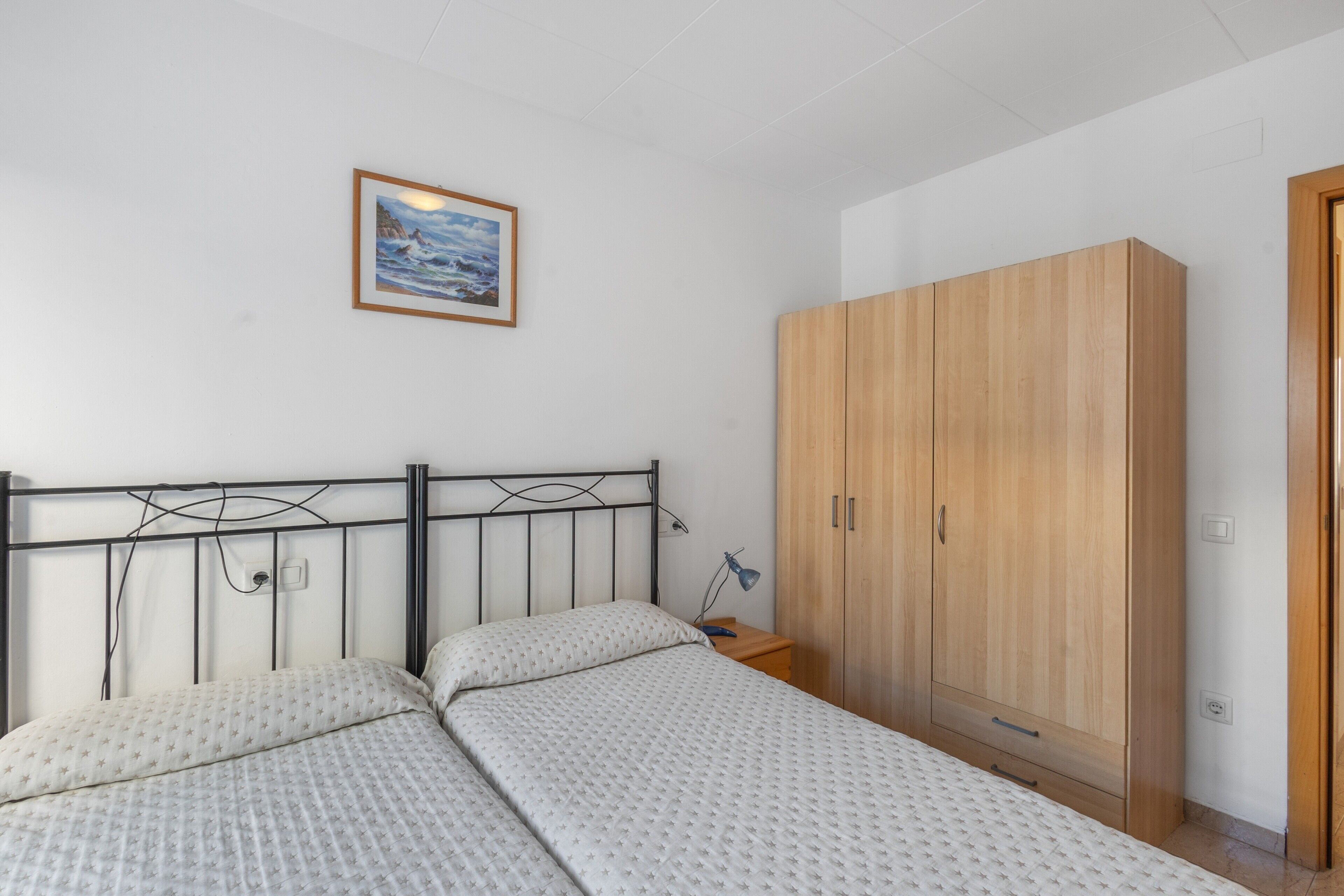 Un dormitorio con cama, armario de madera, mesita de noche, lámpara y un cuadro enmarcado en la pared.
