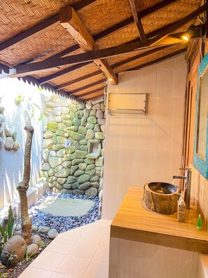 A bathroom with a stone wall, wooden ceiling, and a wooden basin. - Joglo Breeze Guest House and Villa Room 2 (Bali)