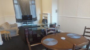 A compact living space with a glass dining table, wooden chairs, a sofa, and a television in the background. - Grace House (Stockton-on-Tees)