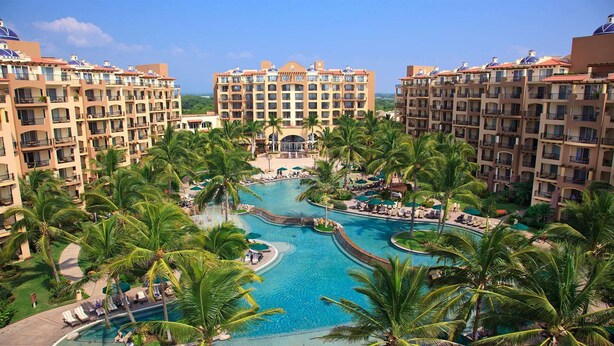 Un resort con varias edificaciones, un área de piscina central rodeada de palmeras y un cielo despejado.