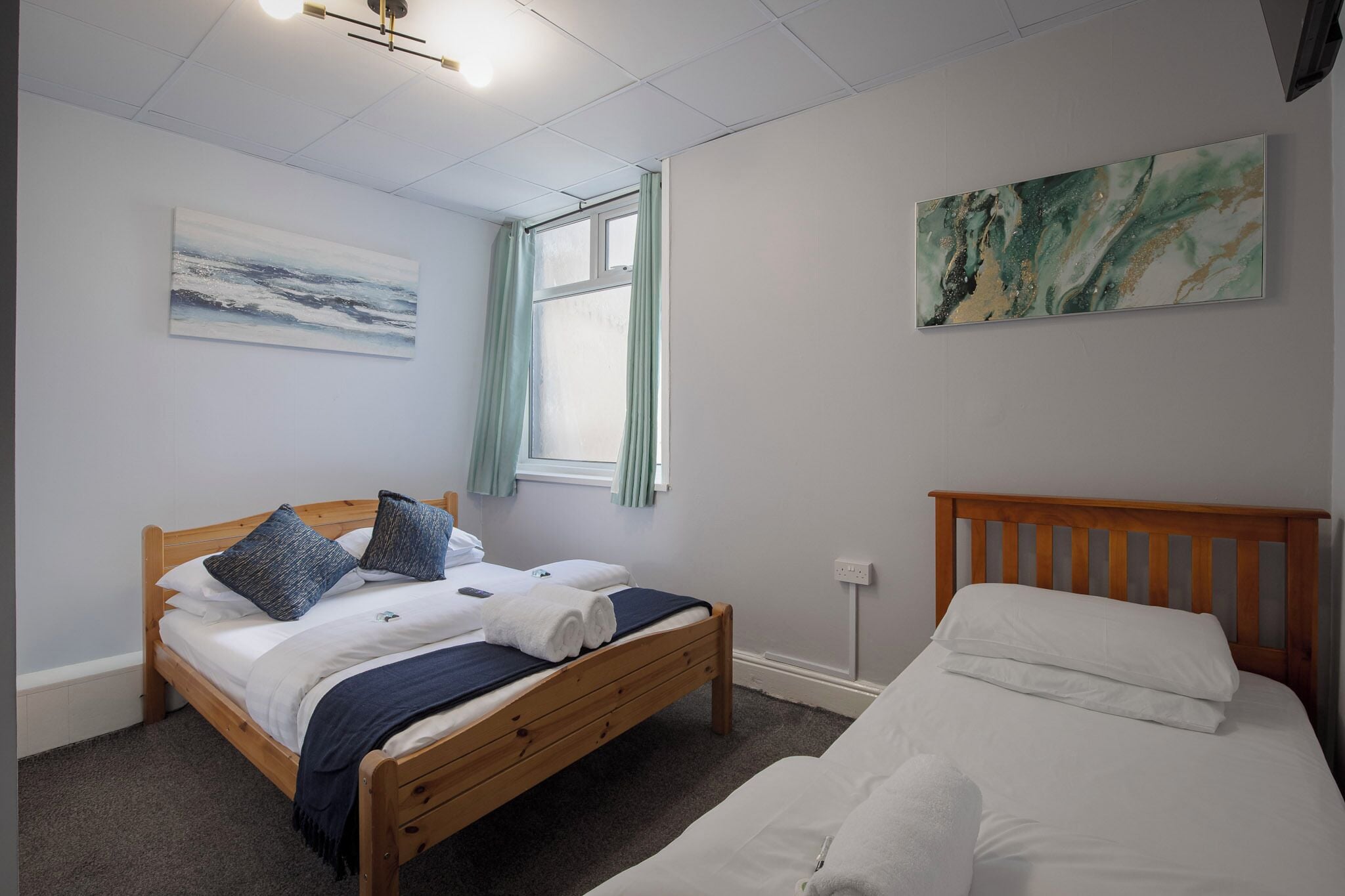 A bedroom with a wooden bed, a smaller bed, a window with curtains, a painting on the wall, and a ceiling light fixture.