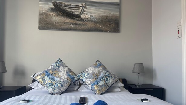 A bedroom with a bed, pillows, a remote control, and a painting of a boat on the wall. - 100 Pipers Hotel (Blackpool)