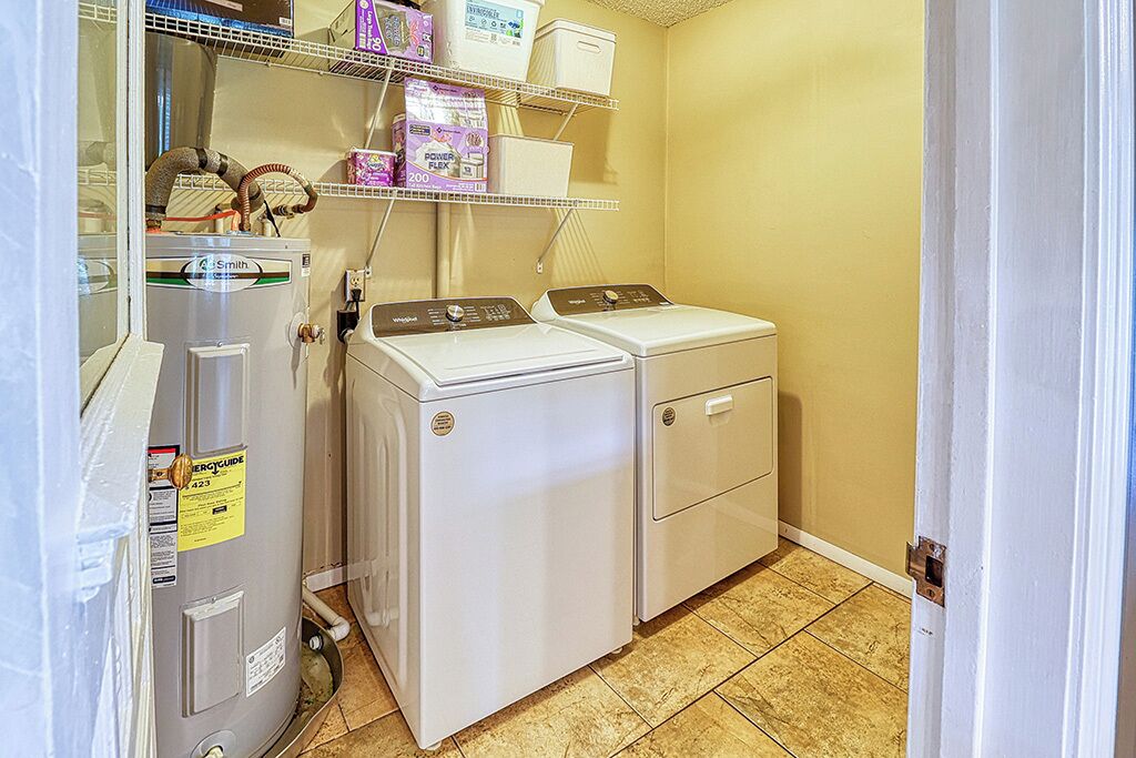 Un cuarto de lavandería con lavadora y secadora de carga frontal, un calentador de agua y estantes con productos de limpieza.