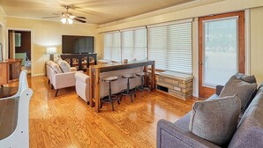 Amplia sala de estar con piso de madera, un conjunto de sofás, un comedor con barra y ventanas grandes con persianas.