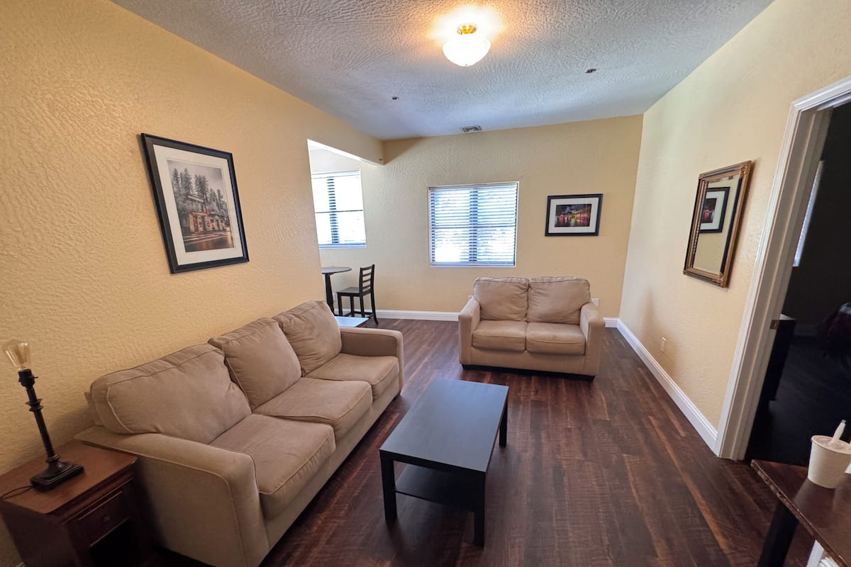 A living room with two beige sofas, a coffee table, and a lamp. There are framed pictures on the walls and a window with blinds.
