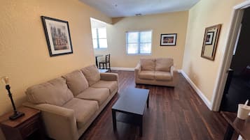 A living room with two beige sofas, a coffee table, and a lamp. There are framed pictures on the walls and a window with blinds.