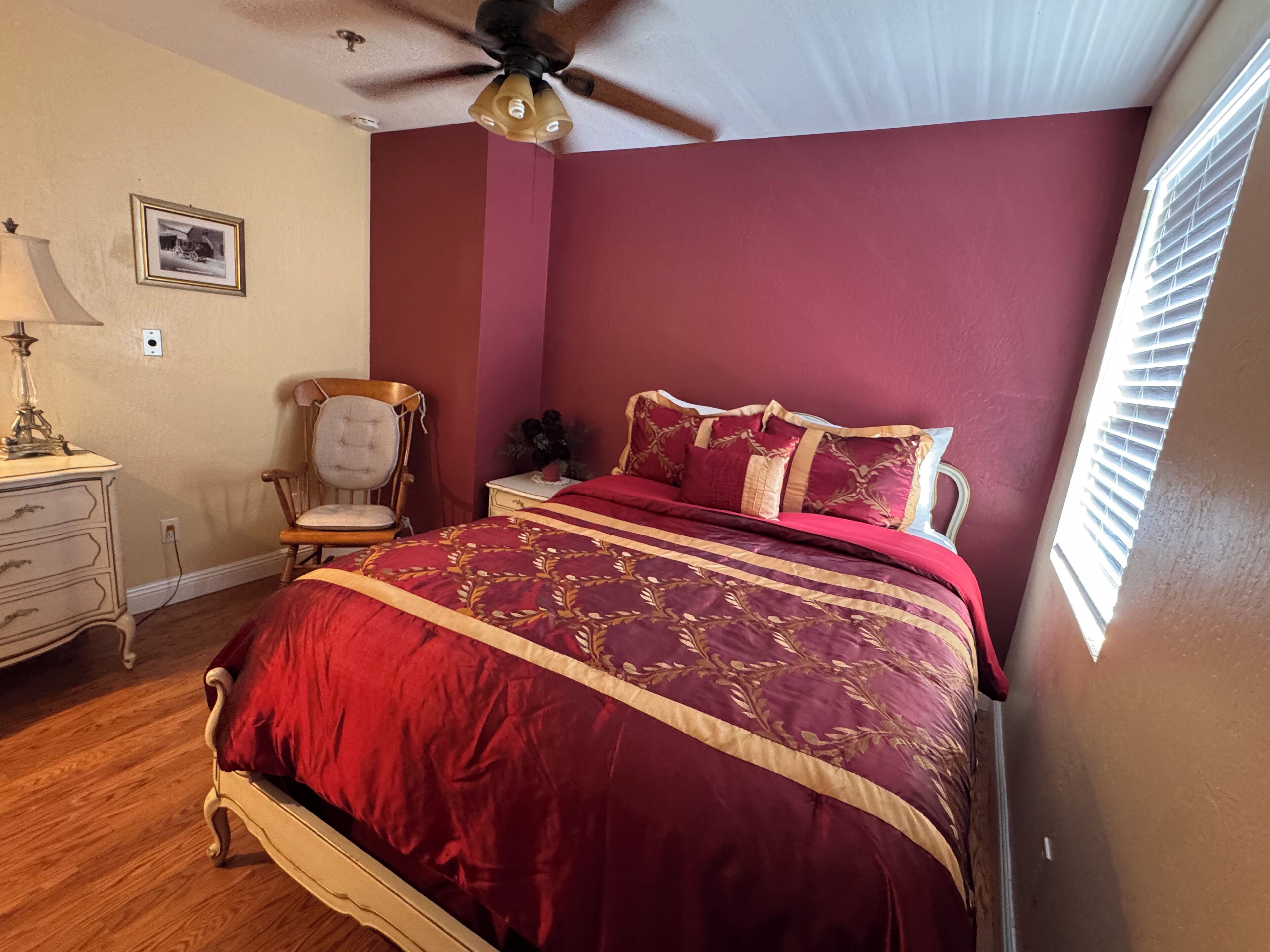 A bedroom with a bed, a chair, a nightstand, a lamp, and a ceiling fan.