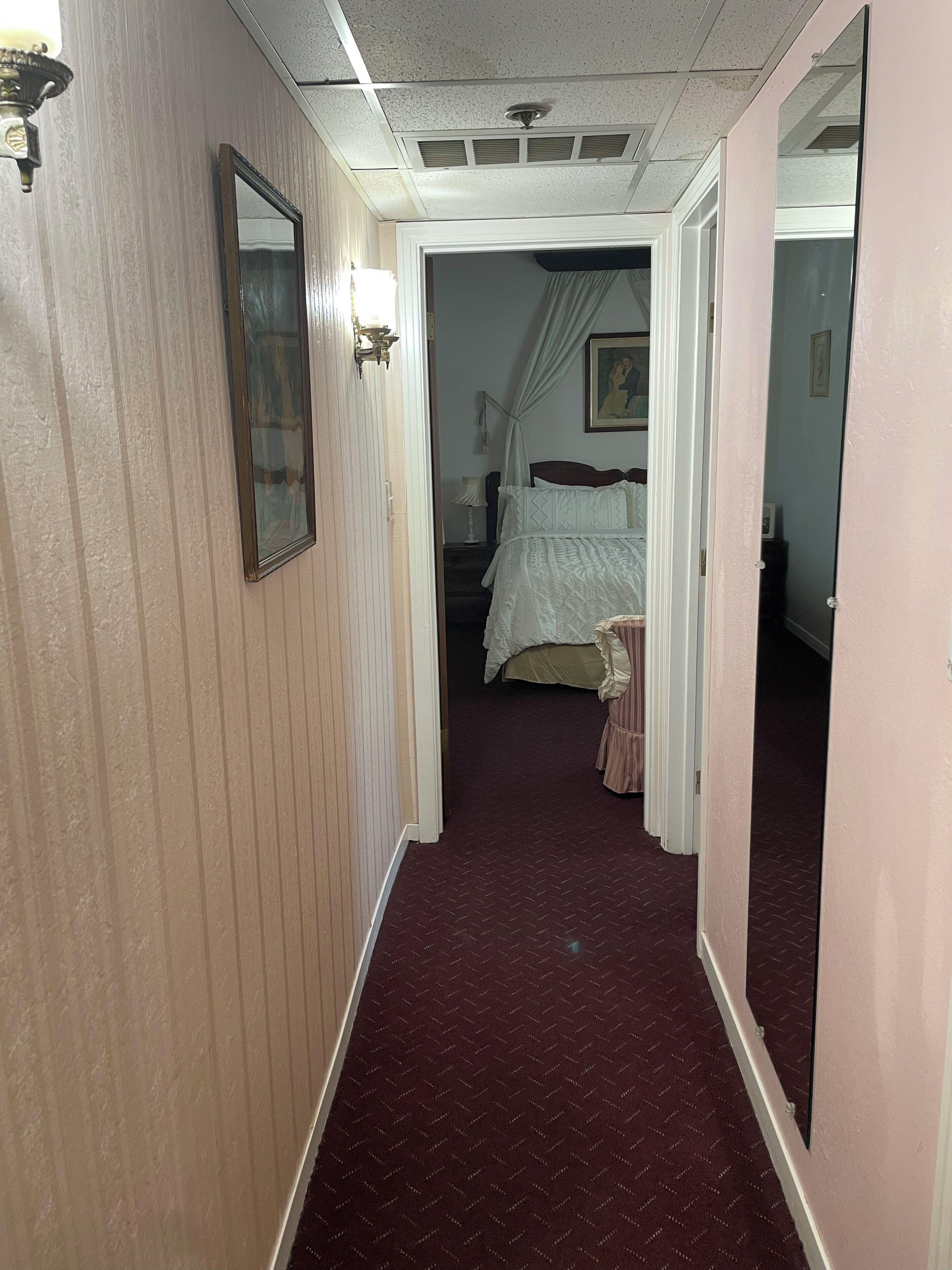 A narrow hallway with a patterned carpet, a bed with a canopy in the background, and a mirror on the right wall.