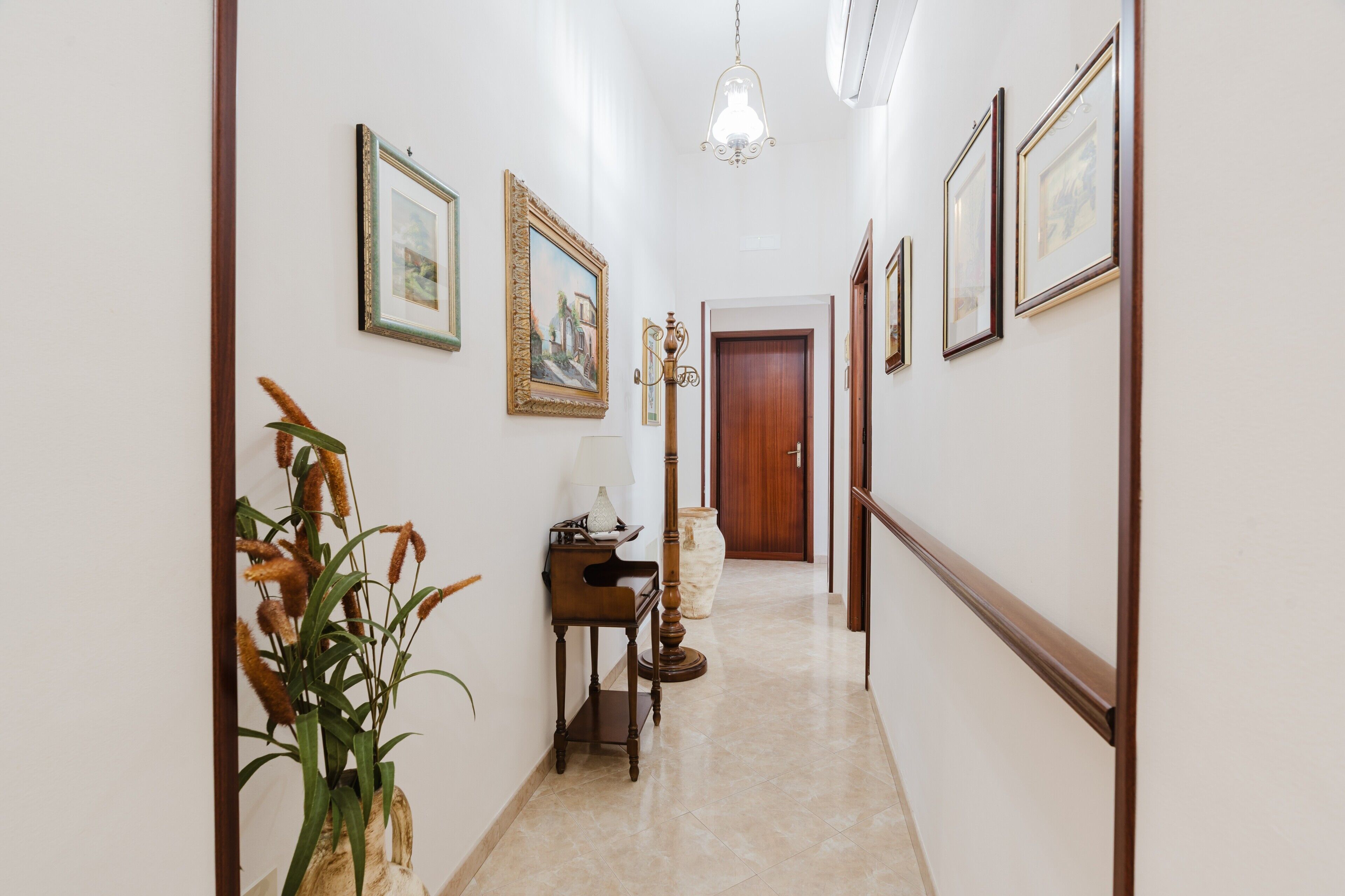 A hallway with framed pictures on the walls, a potted plant, a lamp, and a wooden door at the end.