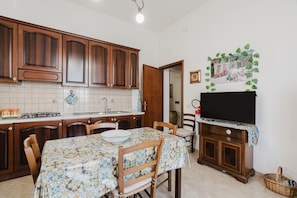 Une cuisine avec des meubles de rangement en bois, un pare-feu en carrelage, une table à manger avec des chaises, un téléviseur posé sur un meuble et un tableau accroché au mur.