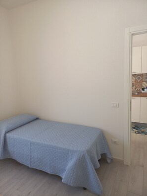 A single bed with a blue bedspread in a room with a wooden floor and a white door leading to a bathroom with a tiled backsplash. - Holiday Home 'Casa Damari-Beachfront' with Sea View and Private Terrace (Petrosino)