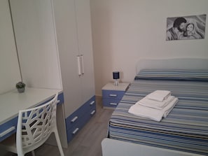 A bedroom with a bed, a desk, a chair, a nightstand, and a wall-mounted lamp.