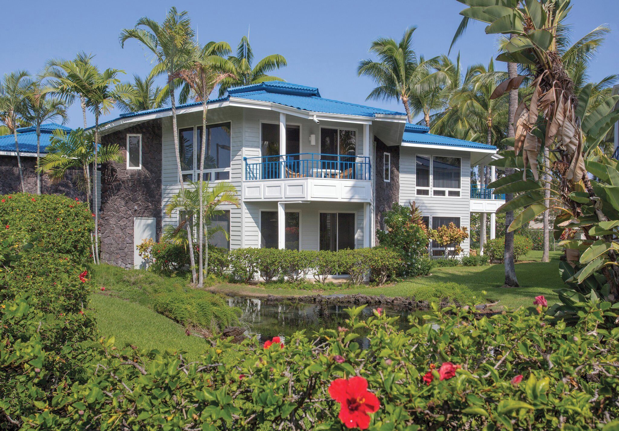 Una casa de dos pisos con techo azul, rodeada de abundante vegetación y palmeras.