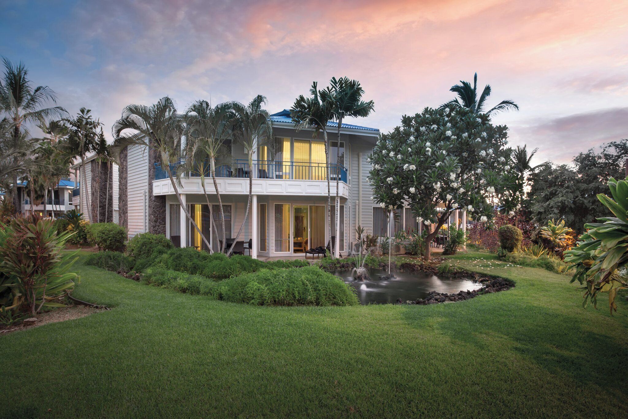A two-story house with a balcony, surrounded by lush greenery and a small pond.
