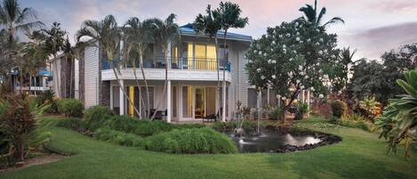 A two-story house with a balcony, surrounded by lush greenery and a small pond.