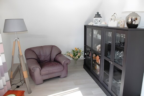 A room with a cushioned armchair, a cabinet with glass doors, a lamp, and a vase with flowers.