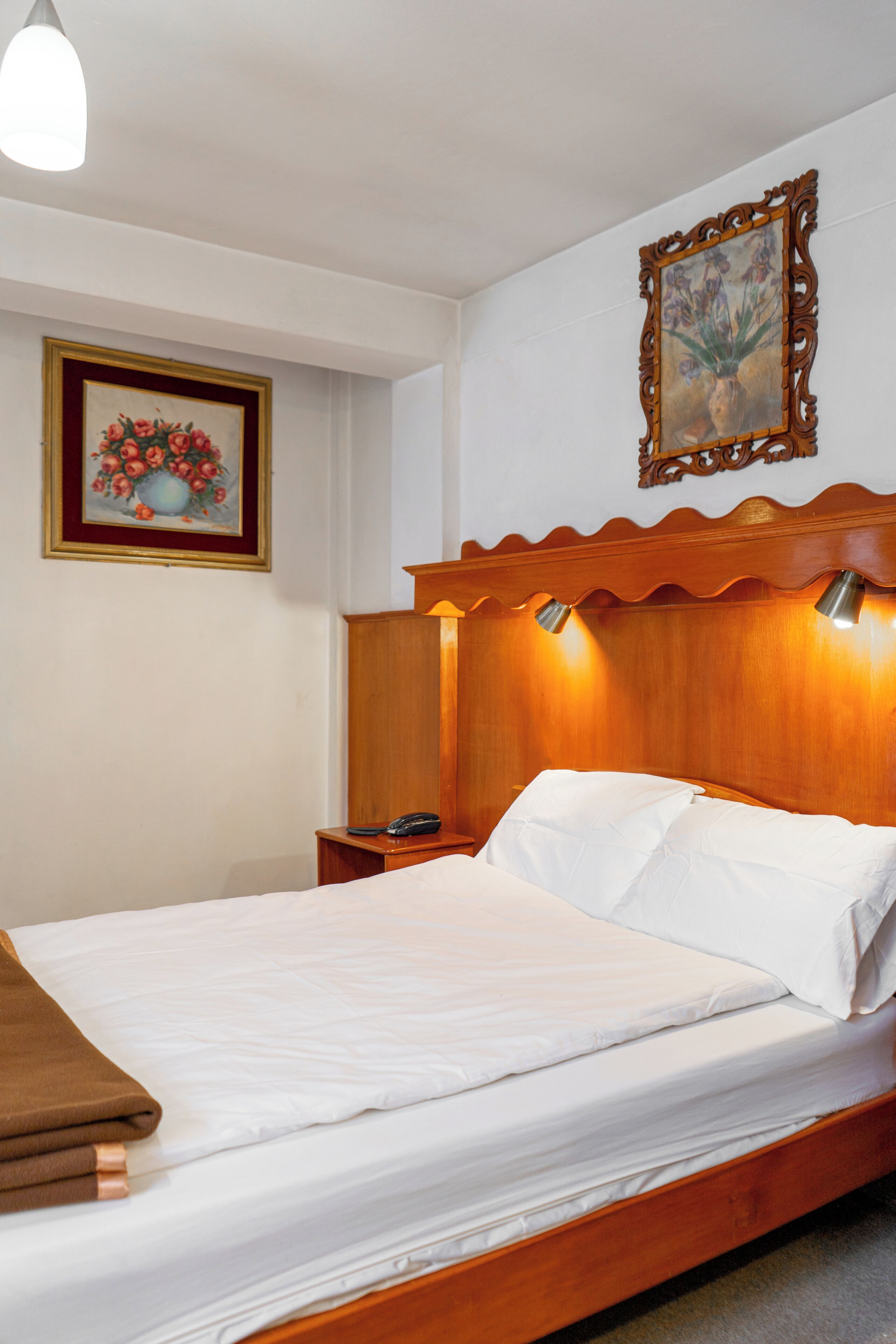 Una cama bien tendida con sábanas blancas, un cabecero de madera con iluminación integrada, una mesita de noche con teléfono y un cuadro de flores enmarcado en la pared.