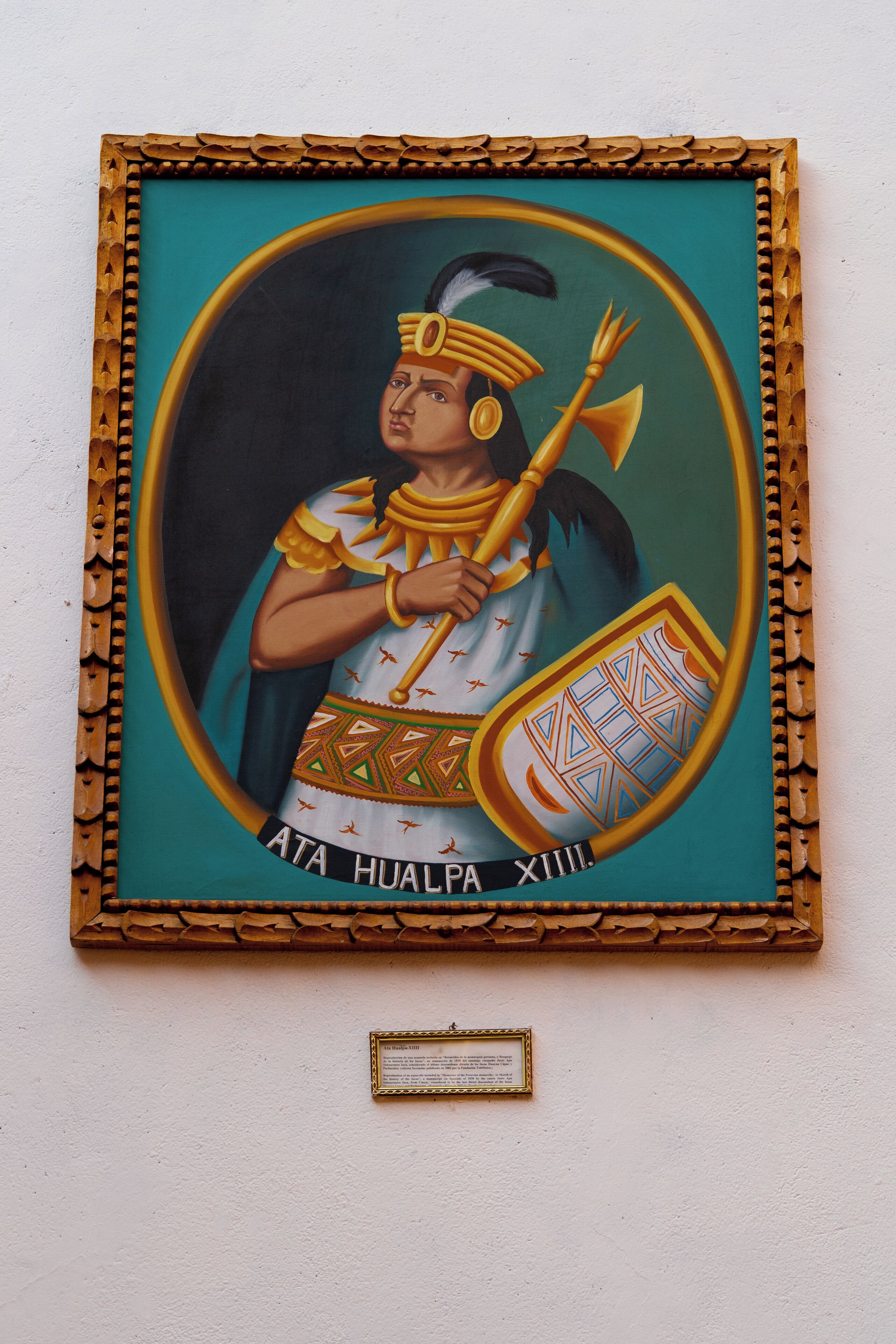 A framed artwork of a figure in traditional attire with a shield and spear, mounted on a wall with a plaque below.