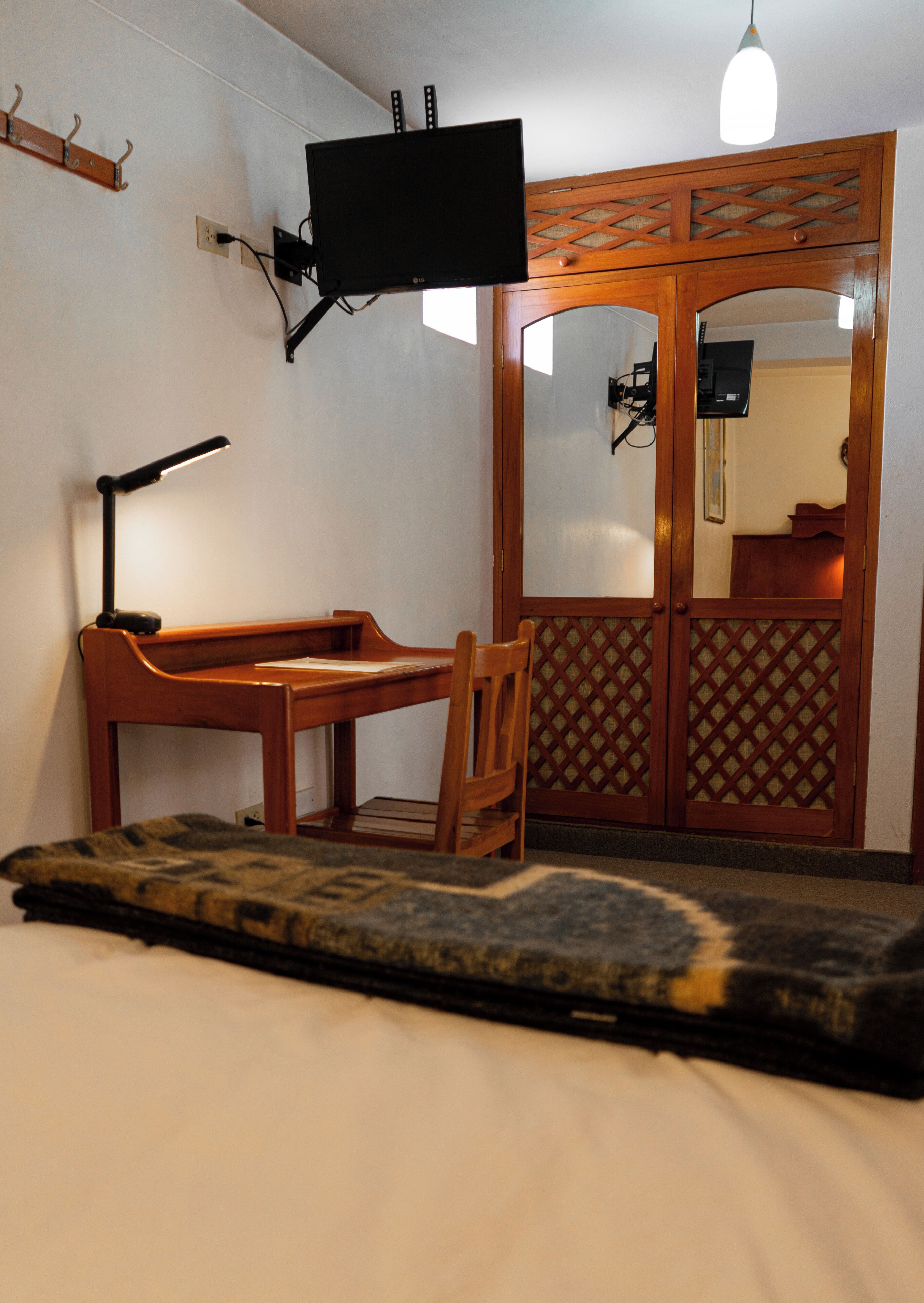 A room with a bed, a desk with a chair, a television mounted on the wall, and a wooden wardrobe with a mirror.
