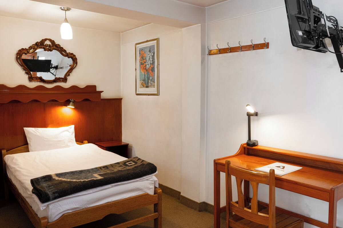 A hotel room with a wooden bed, a desk, a chair, a mirror, a painting, and a television mounted on the wall.