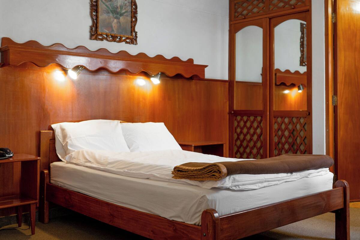 A wooden bed with a headboard, a nightstand, a wall-mounted light fixture, and a wooden door with a lattice design.