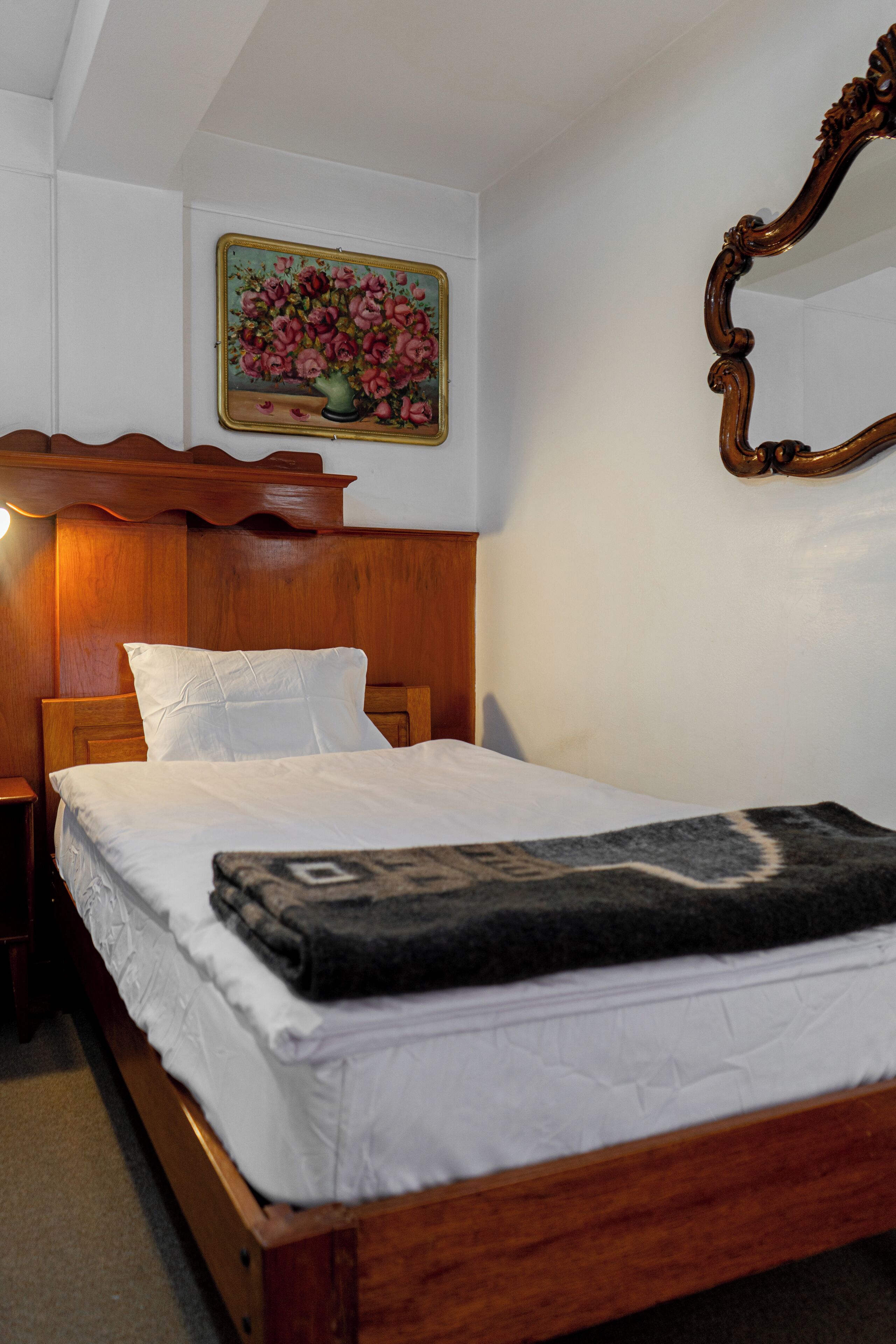 Ein ordentlich gemachtes Bett mit weißen Leinentüchern und einer dunklen Decke, ein hölzerner Kopfteil, ein Nachttisch mit Lampe und ein Blumenbild an der Wand.