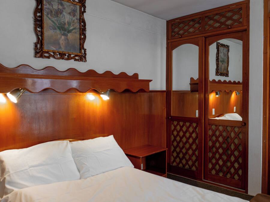 Habitación de hotel con cabecera de madera, cama con ropa de cama blanca y un espejo decorativo con luces.