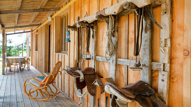 A wooden porch with a rocking chair, a table, and horse tack mounted on the wall.
