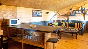 A cozy loft-style room with a wooden ceiling, a kitchenette with a microwave, a colorful striped sofa, and a bookshelf.
