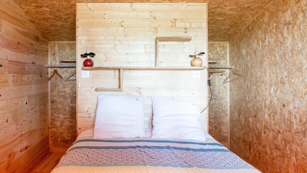 A wooden cabin room with a bed, a shelf with a red object, and a small shelf with a yellow object. - Chalet L'Escale Du Dernier Pin – Peaceful Wooden Retreat in Southwest France (Escalans)