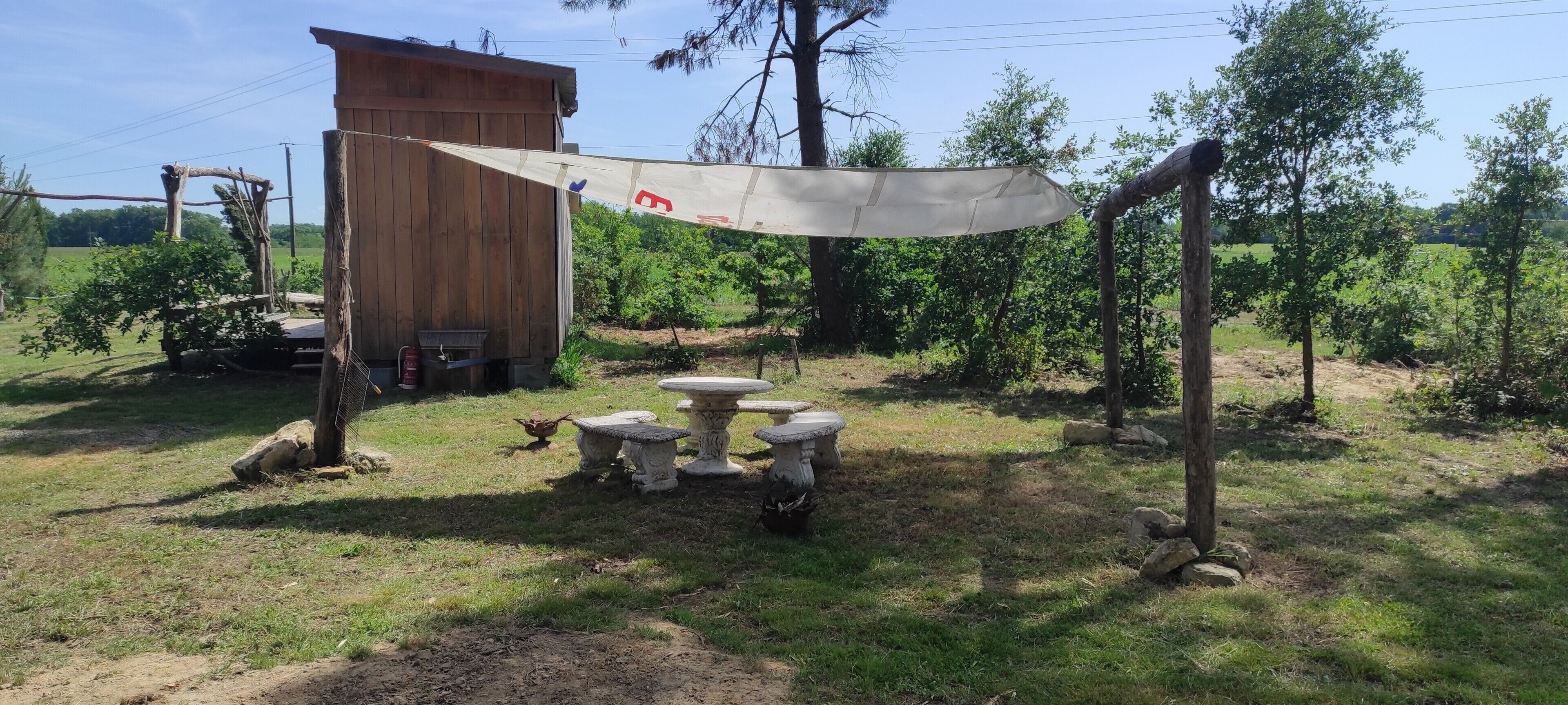 Un espace extérieur rustique agrémenté d’un abri en bois, de tables en pierre et d’un coin ombragé.