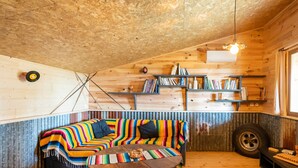 A cozy room with a wooden ceiling, a colorful striped sofa, a round wooden coffee table, and shelves with books and a tire.