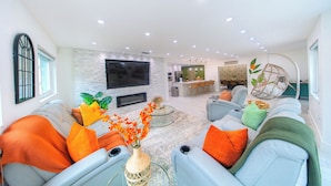 A modern living room with a flat-screen TV, comfortable seating, a fireplace, and a kitchen area in the background.