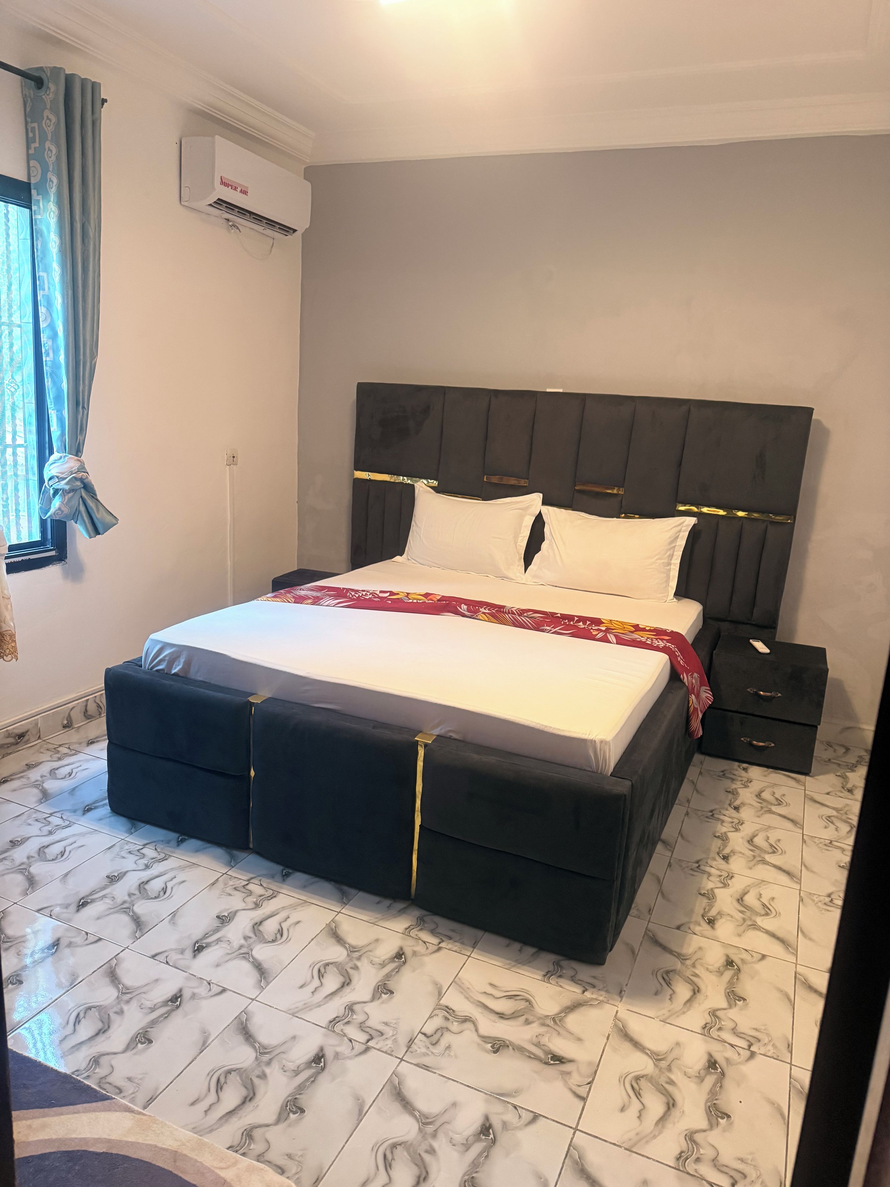 A bedroom with a bed, a nightstand, an air conditioning unit, and patterned floor tiles.