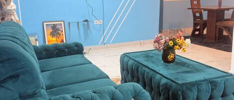 A living room with teal upholstered sofas and ottomans, a flat-screen TV mounted on the wall, and a dining area with wooden furniture.