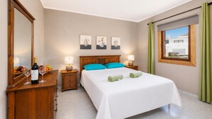 Une chambre à coucher comprenant un lit, deux tables de chevet, un miroir, des œuvres d’art accrochées au mur et une fenêtre avec des rideaux.
