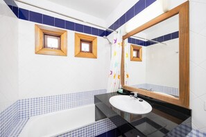 A bathroom with a white sink, black countertop, and a large mirror. There are two small windows with wooden frames on the wall, and a shower curtain with colorful patterns.