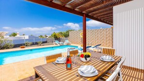 Un espace repas aménagé au bord de la piscine, avec une table en bois dressée pour deux personnes, une bouteille de champagne Duet et un panier de fruits.