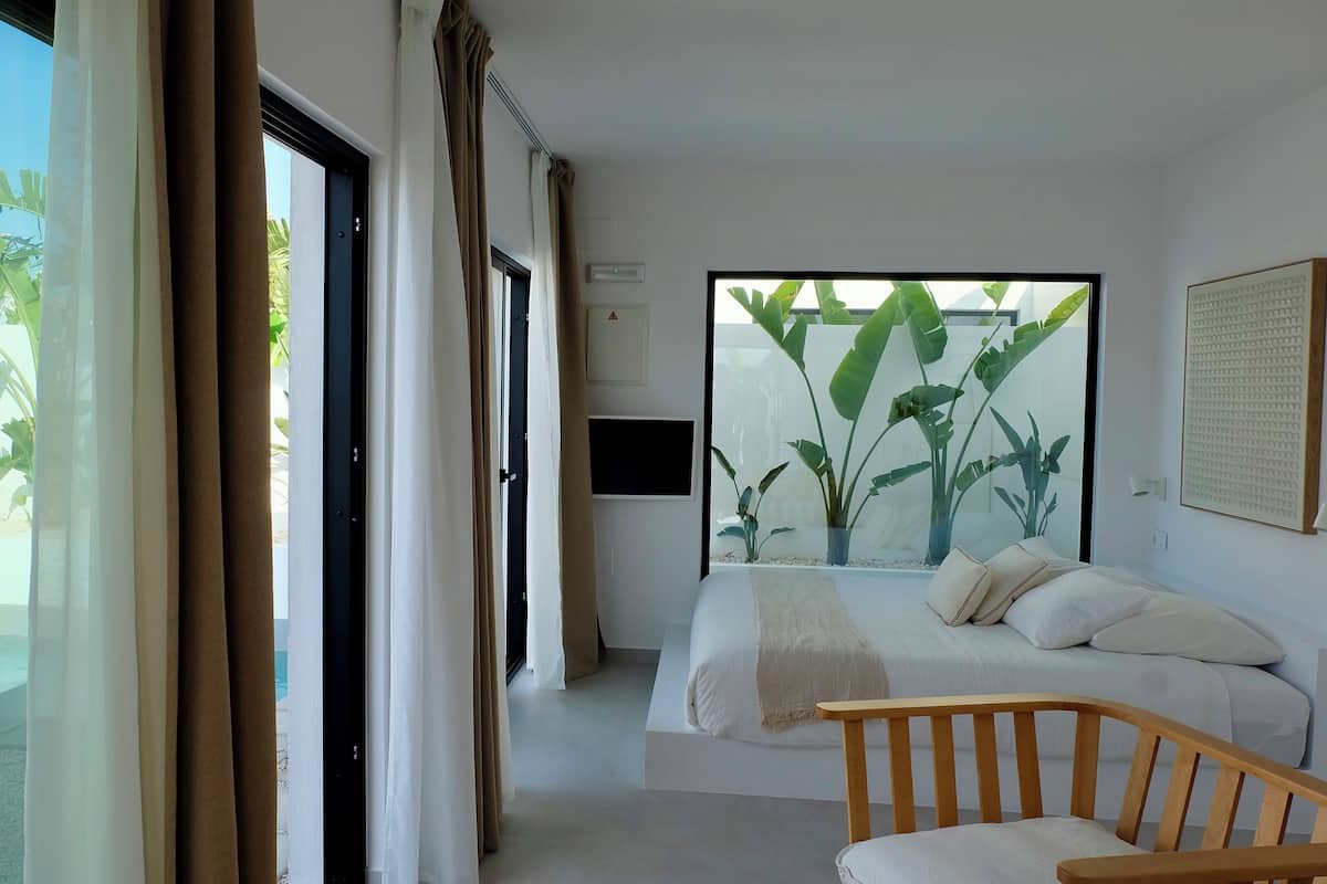 A modern hotel room with a large bed, a wooden chair, and a view of the outdoors through large windows.